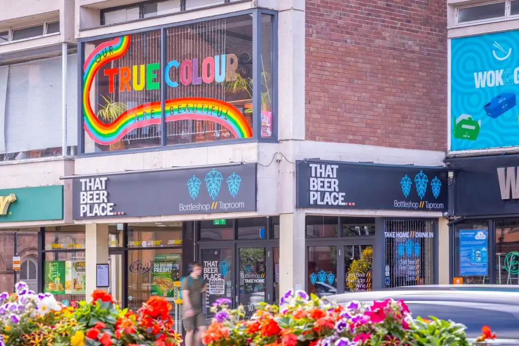 Bottleshop That Beer Place beneath a rainbow window reading True Colours Are Beautiful, with bright flowers in the foreground.
