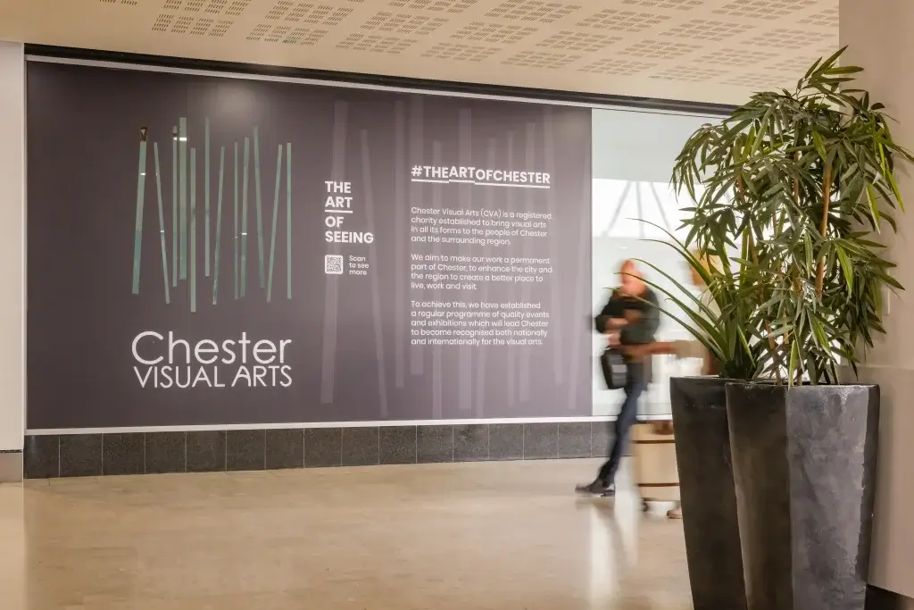 A man walks past a wall display for Chester Visual Arts, promoting local arts and events, with greenery in the foreground.