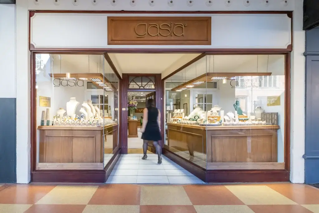 A woman in a black dress enters a jewellery shop named "gasia," showcasing elegant displays of necklaces and rings in its windows.