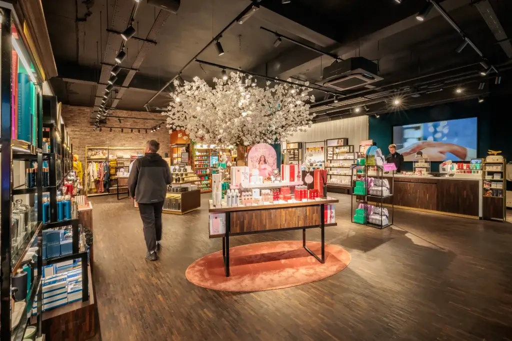 Boutique interior with central display under a large white blossom tree, cosmetic shelves and a customer.
