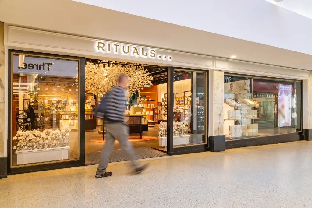 A blurred figure walks past the inviting entrance of a Rituals store, showcasing shelves filled with beauty products and decorative blossoms.