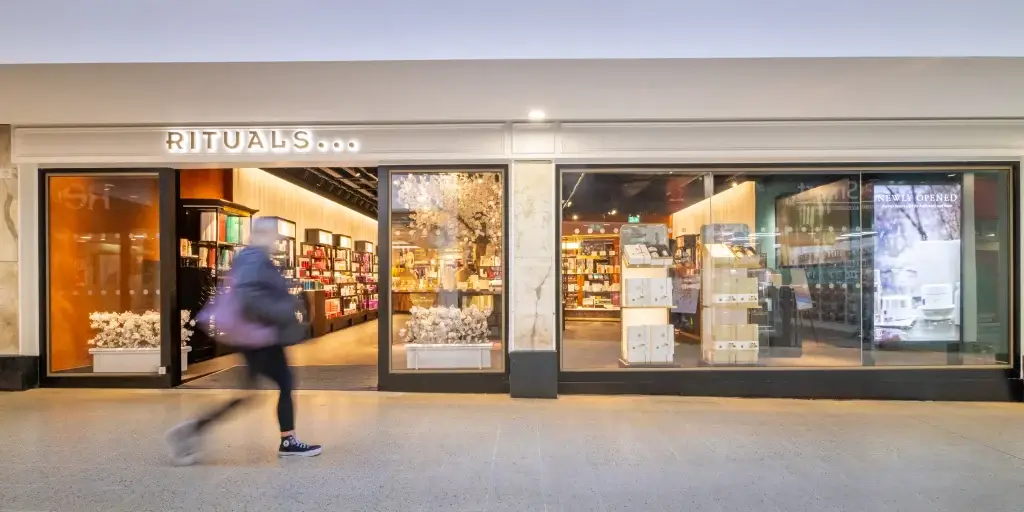A blurred figure walks past the inviting entrance of a Rituals store, showcasing shelves filled with beauty products and decorative blossoms.