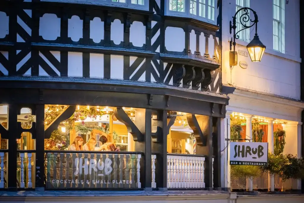 A cozy, illuminated restaurant named "Shrub," featuring outdoor seating and a warm ambiance, set in a charming historic building.