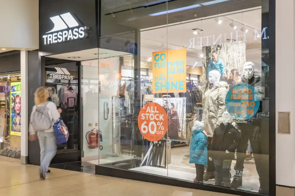 A shopper approaches the Trespass store entrance featuring a promotional sign for 60% off all stock, with outdoor gear displayed inside.