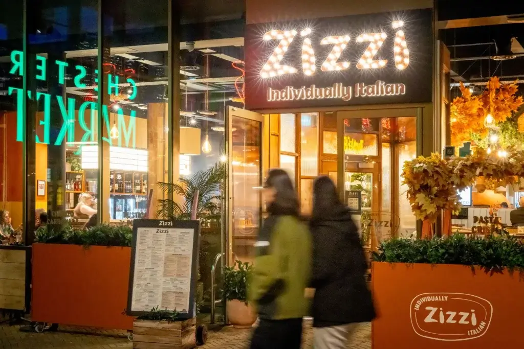 A vibrant restaurant entrance featuring "Zizzi" sign, with patrons inside, surrounded by plants and autumn decor. Two people walk by.