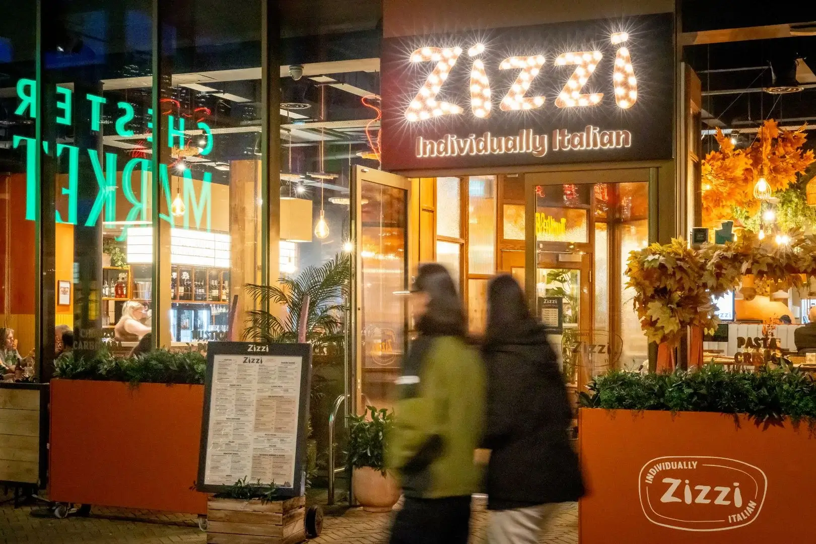 Zizzi restaurant entrance at night with lit marquee reading Individually Italian, menu board, orange autumn decor and two blurred passersby.