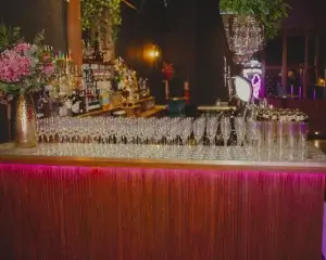 Bar counter lined with dozens of empty champagne flutes, bottles and beer taps behind, pink flowers and magenta lighting.