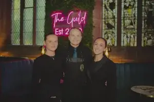 Three smiling staff in black, centre wearing an apron, standing before a neon Guild sign and stained-glass windows.