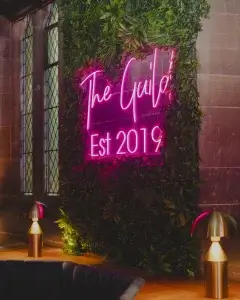 Pink neon sign saying The Guild Est 2019 mounted on a living green wall, flanked by brass lamps and a stained-glass window.