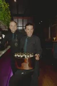 Two bar staff in black shirts and teal ties, woman excitedly holding a copper bucket filled with bottled beers.