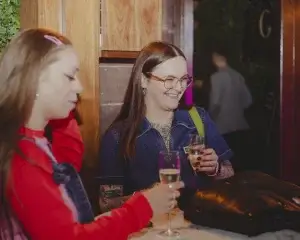 Two women at a bar laughing and holding glasses of sparkling wine; woman on right wears glasses, denim top and visible tattoos.