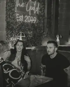 Three people in a dim lounge booth, a smiling woman and bearded man talking over champagne under a neon Est 2019 sign.