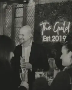 Smiling bald man in a suit holding a drink at a social gathering with a neon sign reading The Guild Est 2019 behind him.