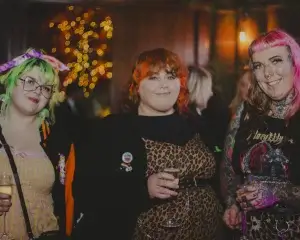 Three women at a party with colourful hair (green, orange, pink), tattoos and glasses, wearing leopard print and holding champagne flutes before warm bokeh lights.