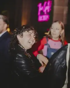 Person with curly hair in a black leather jacket laughing next to a friend in red and denim, pink neon sign blurred behind.