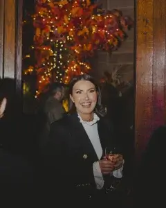 Smiling woman in a black blazer and ruffled white blouse holding a wine glass, fairy lights and autumnal foliage behind her.