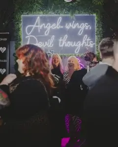 Two women laughing in front of a neon sign reading Angel wings, Devil thoughts at a crowded night event