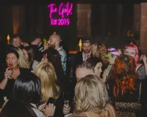 Crowded indoor party with people mingling under a pink neon sign reading Est 2019, guests holding drinks.