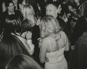 Black-and-white crowded party scene with a woman in a white strappy dress from behind and a man holding a champagne glass.