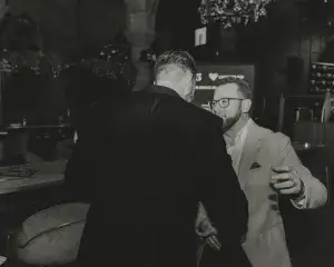 Bearded man in glasses and light blazer reaching to embrace a man in a dark suit at a dim indoor event.