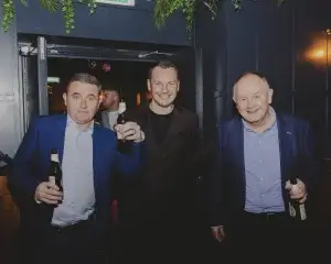 Three men in jackets smiling and holding beer bottles in a dim bar doorway, plants hanging above.