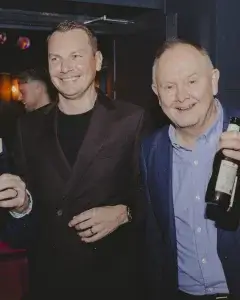 Two smiling men in blazers at a dimly lit social gathering, one holding a beer bottle and the other in a dark suit.