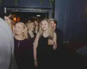 Young blonde woman in a black dress smiling and holding a yellow drink with a straw in a dim bar, people around her