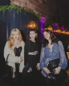 Three women in a dim venue with warm lights and greenery: blonde in cream, centre in black, right in blue floral dress.