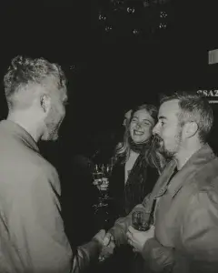 Three people at a black-and-white party scene; two men shake hands while a woman laughs holding two champagne flutes.