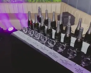 Row of champagne bottles and heart-shaped glass trophies on a table with handwritten award labels, purple uplighting.