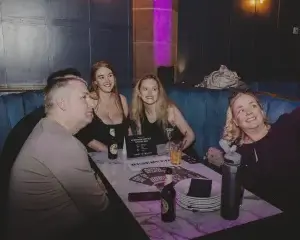 Four adults smiling in a dim lounge booth, one woman taking a selfie, drinks and a Reserved sign on the table.