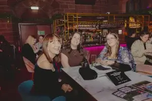 Three women smiling at a marble table in a bar, drinks and purses in front of them and a backlit shelf of bottles behind.