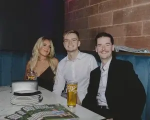 Three young adults—a blonde woman and two men—smile in a booth with drinks and stacked plates on a marble table.
