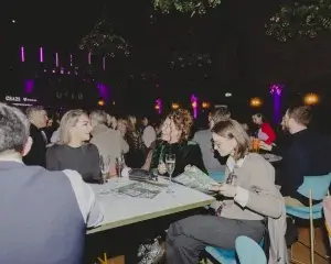 Group at a table in a dim purple-lit venue, three women chatting while one reads a programme with a glass of champagne.