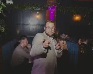 Bearded man in glasses and light jacket pointing at the camera inside a dimly lit bar booth with seated patrons behind
