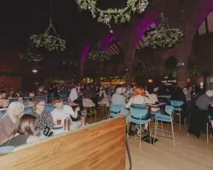 Crowded bar in a converted church with high arches, purple uplighting, hanging greenery chandeliers and patrons at tables.