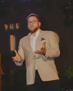 Man in beige blazer and glasses speaking on stage with a headset mic, animatedly gesturing with both hands.