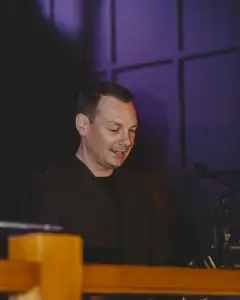 Man in a dark suit smiling behind a wooden podium with a microphone against a purple-lit panelled background.