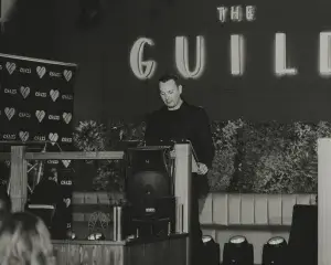 Man in dark suit speaking at a podium on a stage with a lit THE GUILD sign and CSA25 heart backdrop.
