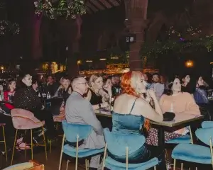 People seated in a dim bar; foreground red-haired woman in teal satin dress at a table with blue velvet chairs.