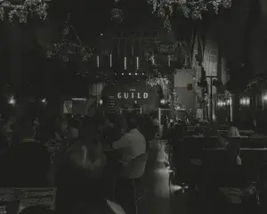 Crowded dimly lit venue with chandeliers and gothic arches, audience seated facing a stage lit with a sign reading THE GUILD