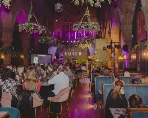 Crowded vaulted hall labelled GUILD with seated diners facing a purple-lit stage, hanging greenery and disco balls.