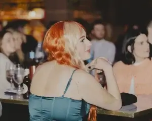 Woman with half-orange, half-blonde wavy hair in a teal satin dress seated at a table in a dimly lit venue, hands clasped.