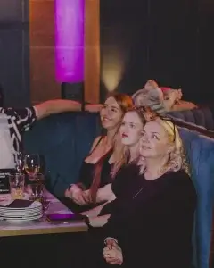 Three women seated on a blue booth in a dimly lit lounge, smiling and looking up, with drinks and plates on the table.
