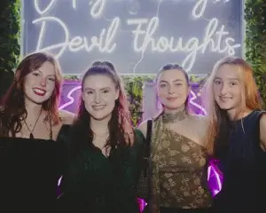 Four young women smiling together in front of a neon "Devil thoughts" sign with a leafy backdrop.