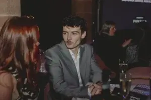 Young man in a grey suit with a moustache talking to a red-haired woman at a table with champagne glasses.
