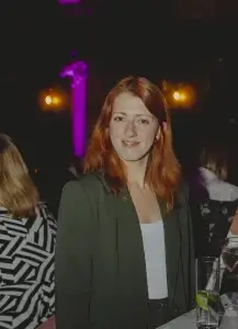 Red-haired woman in a dark blazer and white top smiling at the camera in a dimly lit event space with drinks on the table.