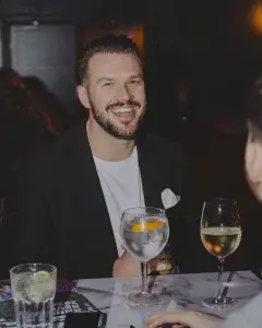 Smiling bearded man in a black blazer and white T-shirt with a large cocktail garnished with orange and a glass of white wine.