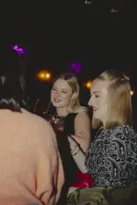 Two young women smiling at a night gathering, one holding a wine glass and the other wearing a patterned top with nose ring.