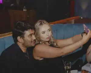 Blonde woman taking a selfie with a dark-haired man leaning in at a blue booth, champagne flute and plates visible on the table.
